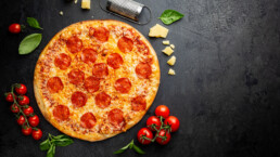 Velsmakende pepperoni pizza og matlagingsingredienser tomater basilikum på svart betongbakgrunn. Topputsikt av varm pepperoni pizza.